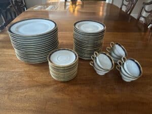 Stacks of white china plates with gold edges and several teacups with saucers on a wooden table.