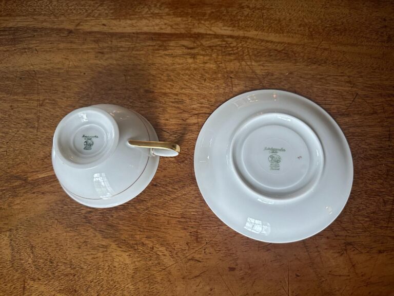 A white porcelain teacup set upside down on a saucer with visible markings, next to an empty saucer on a wooden table.