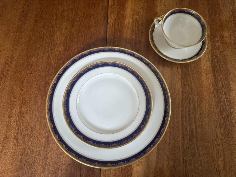 A set of elegant china with blue and gold trim is arranged on a wooden table, including a dinner plate, salad plate, bread plate, cup, and saucer.