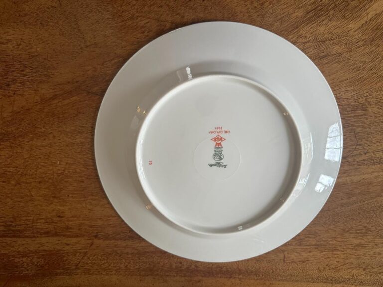 A white ceramic plate upside down on a wooden surface, featuring a red and green emblem with text on the bottom.