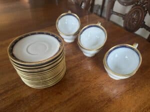 A stack of white saucers next to three teacups with blue and gold trim on a wooden table.