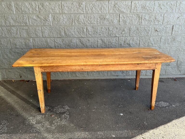 An old wooden table with a rectangular top and four legs stands on a concrete floor against a gray brick wall.