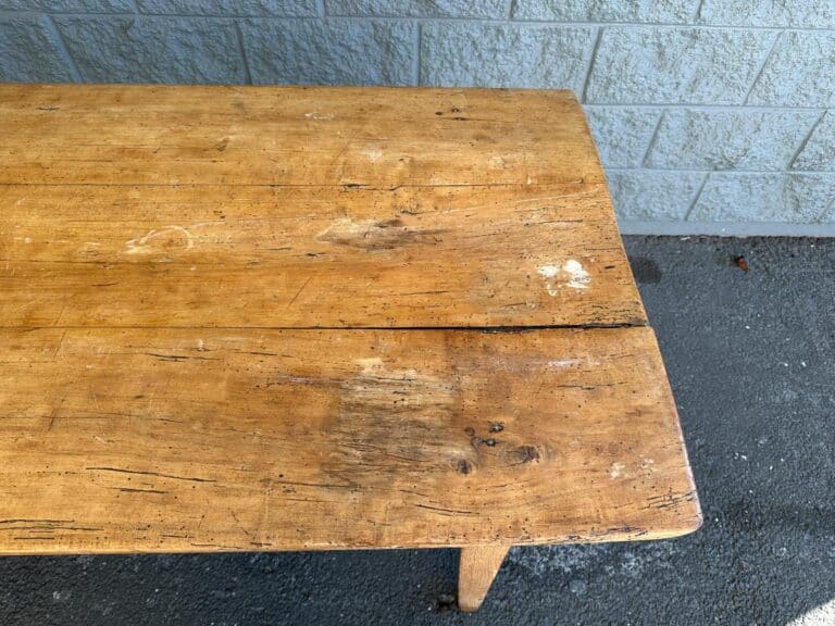 A worn wooden table with visible scratches and marks stands on a concrete surface near a light-colored brick wall.