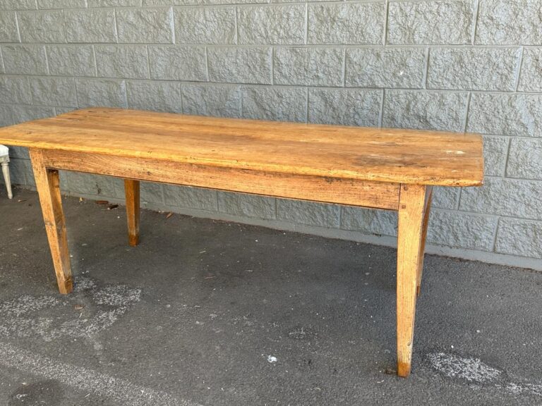 A long, rectangular wooden table with four legs stands on a rough concrete surface against a gray brick wall.