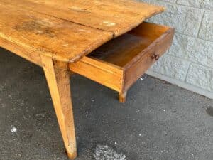 An old wooden table with visible wear shows an open drawer. The table is positioned on a concrete floor near a stone wall.