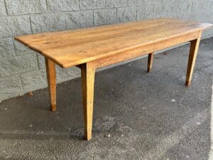 A wooden table with a rectangular top and four tapered legs stands on a concrete surface near a stone wall.