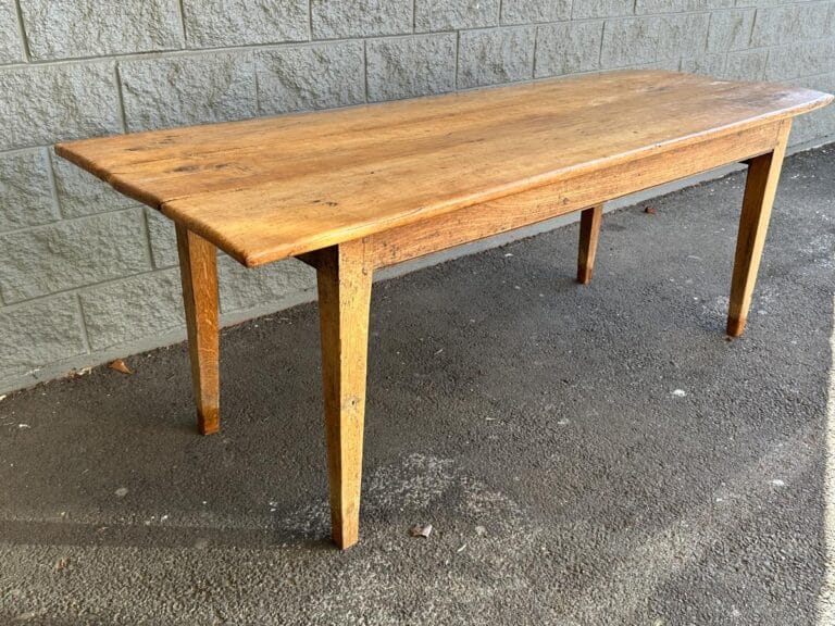 A wooden table with a rectangular top and four tapered legs stands on a concrete surface near a stone wall.