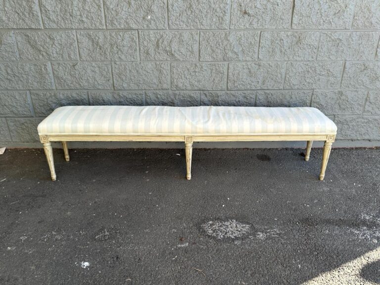 A striped bench with a light-colored cushion is placed against a textured gray wall on a paved surface.