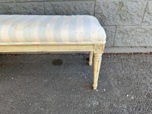 Wooden bench with light blue and white striped upholstery, placed against a stone wall on a paved surface.