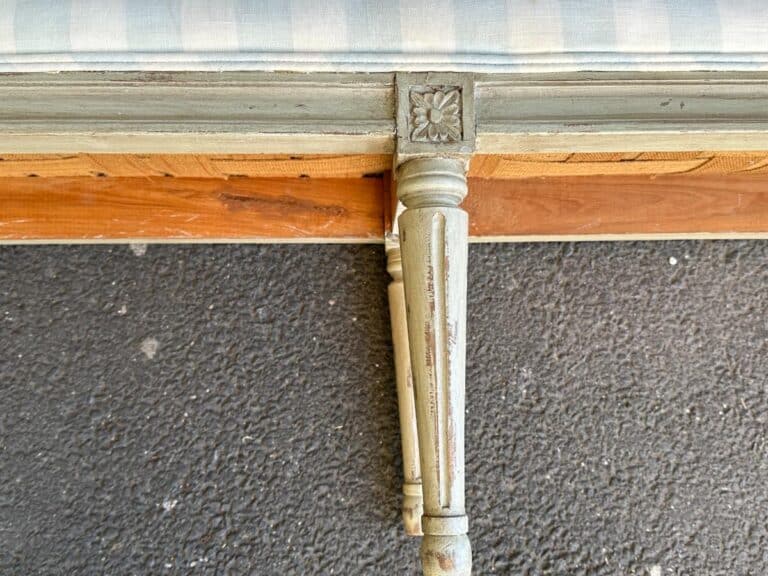 Close-up of a vintage wooden chair leg with decorative carving, attached to a partially visible seat with a striped cushion, on a textured dark surface.