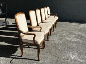 Six beige upholstered wooden chairs are lined up outdoors on a concrete surface in sunlight, against a dark wall.