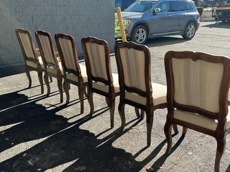 Five wooden chairs with cream fabric backs are lined up outdoors on a sunny day, casting long shadows on the pavement. A gray vehicle is parked nearby.