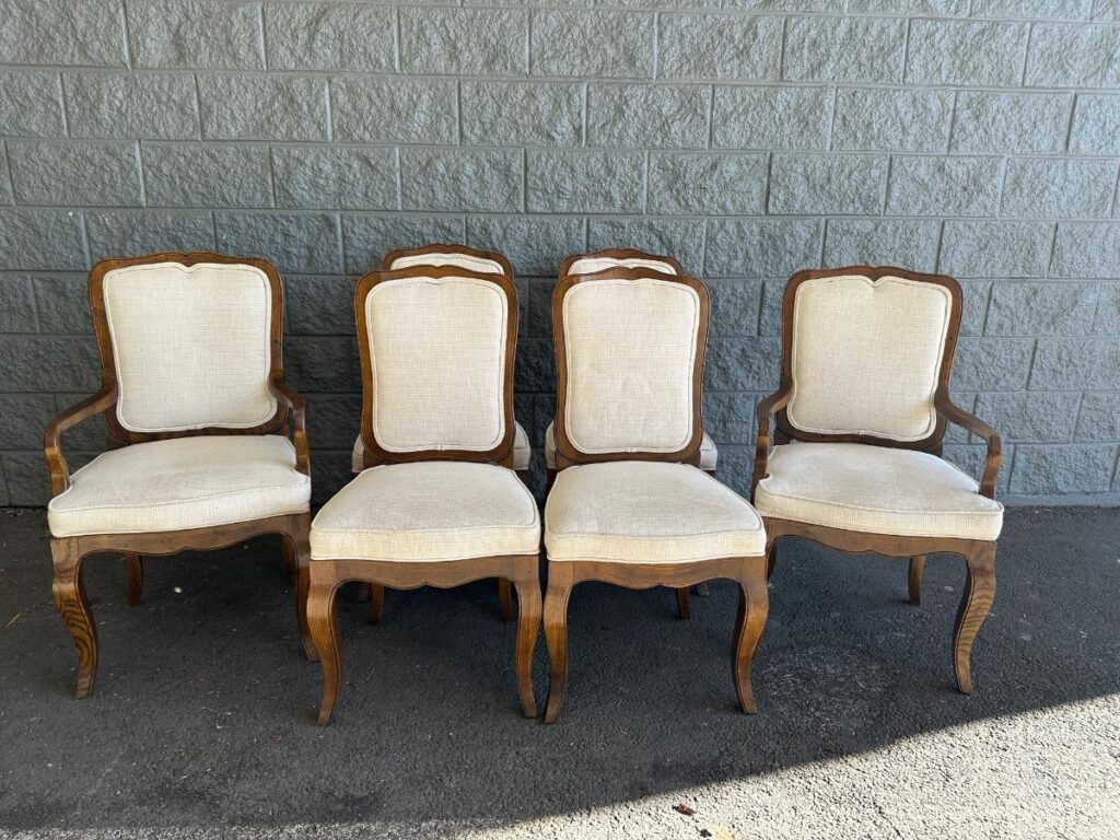 Four wooden chairs with beige upholstered seats and backs, lined up against a gray brick wall outdoors.