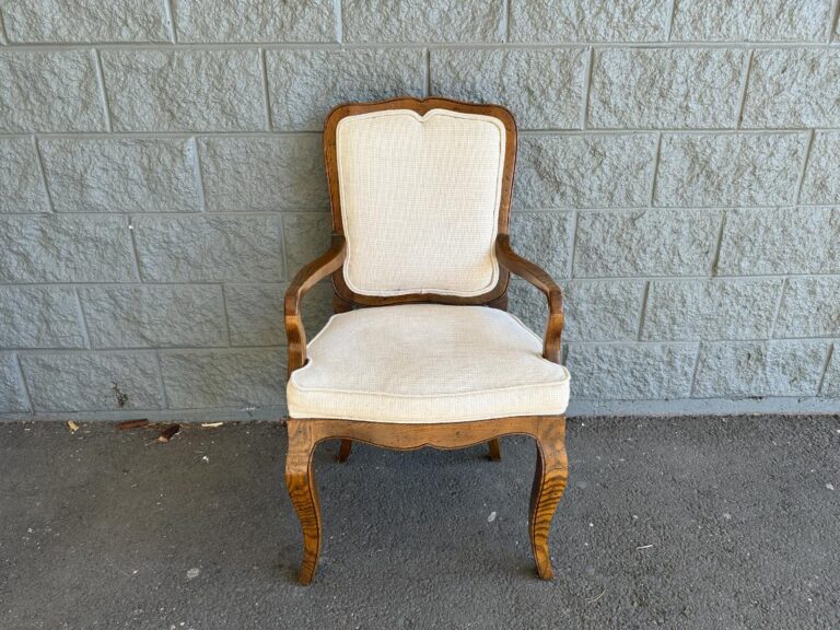 A vintage wooden armchair with a cushioned seat and backrest is placed against a gray brick wall on a concrete floor.