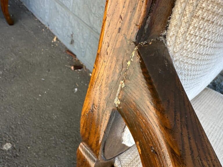 Close-up of a wooden chair with visible cracks on the armrest and exposed joints, set against a concrete floor.