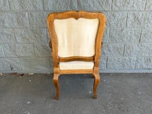 A wooden chair with a light-colored upholstered backrest is positioned against a gray brick wall on a concrete floor.