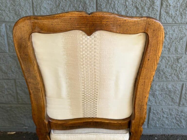 A wooden chair backrest with a carved frame and light fabric upholstery against a gray brick wall.