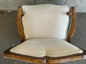 Wooden chair with curved armrests and light beige cushioned upholstery, placed on a paved surface.