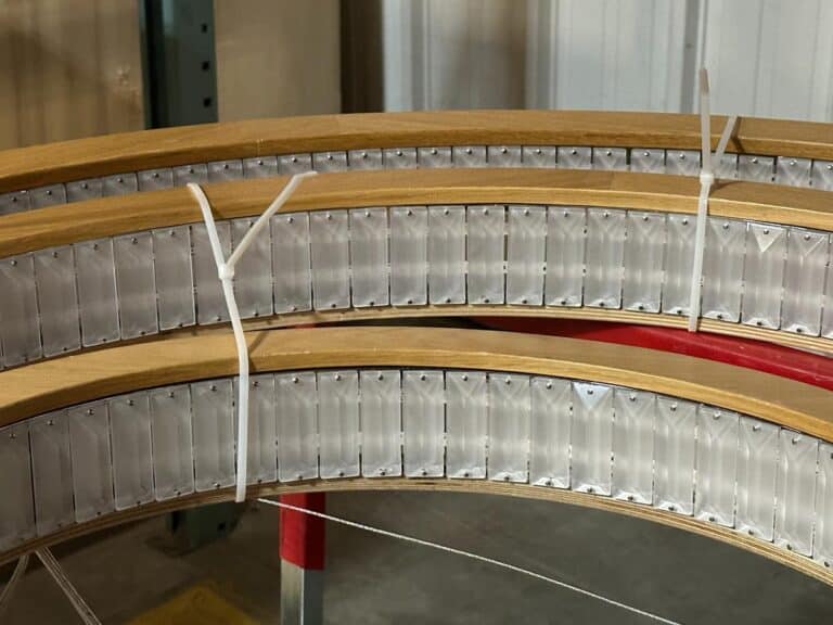 Close-up of two curved wooden beams with attached metal brackets, secured with white zip ties. Background includes a metal shelf and a gray wall.