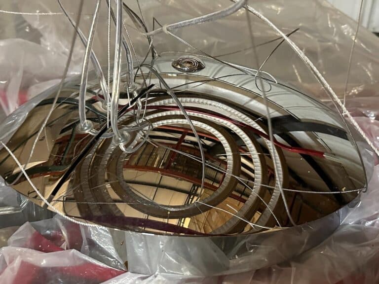 Close-up of a reflective metallic sculpture with intricate wire designs, resting on a plastic sheet. The surface reflects surrounding structures in a distorted manner.