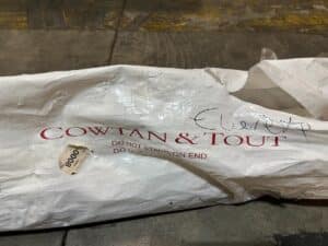 A crumpled white package labeled "COWTAN & TOUT" lies on the ground, with "DO NOT STAND ON END" printed below. A handwritten note and a small sticker with "8000" are visible.