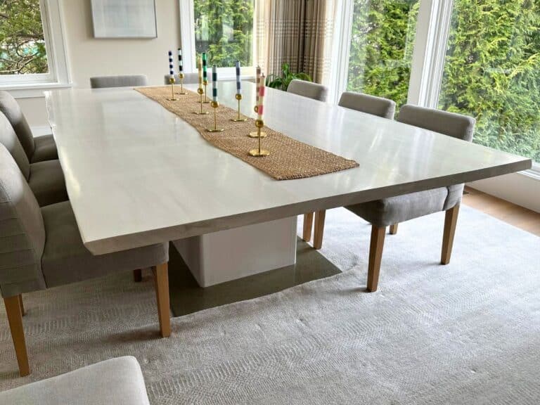 A modern dining room features a Live Edge Custom Dining Table from Cocoon, surrounded by gray upholstered chairs and accented with a woven runner and decorative candlesticks. Large windows reveal the lush greenery outside.