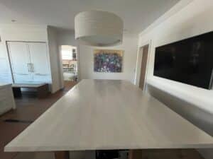 A long, light-colored wooden table under a pendant light in a bright room with white cabinets, a painting, and a wall-mounted TV.