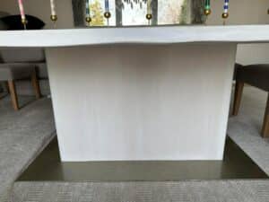 A square, white table with a solid base and decorative candles on top, situated in a carpeted room with chairs around it.