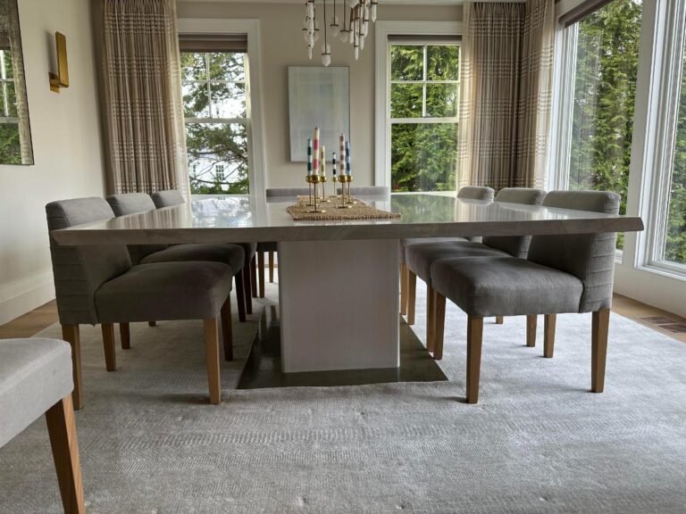 Modern dining room with a large rectangular table surrounded by eight gray chairs. Windows offer a view of greenery outside. A central chandelier and colorful candles are above the table.