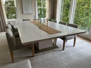 A large dining table with eight gray upholstered chairs, a woven table runner, and brass candle holders sits in a bright room with large windows and a view of greenery.