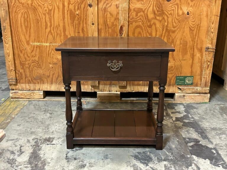 A wooden side table with a single drawer and lower shelf, set against a wooden backdrop.