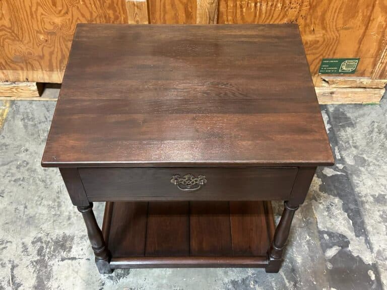 Dark wood side table with a single drawer featuring an ornate metal pull, and a lower shelf. Placed on a concrete floor, wooden panels are visible in the background.