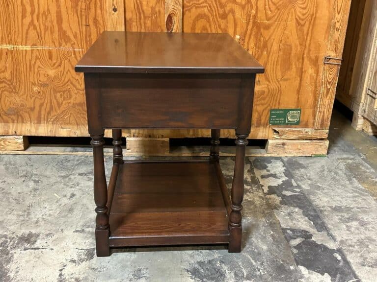 A wooden side table with a lower shelf, set against a wooden crate backdrop on a concrete floor.