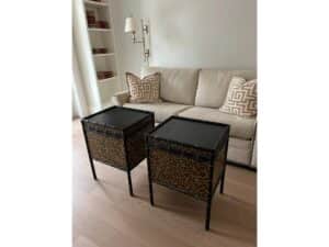 Two leopard print square tables with black tops are placed in front of a beige sofa. The sofa has two patterned cushions and is flanked by bookshelves and a wall lamp in the background.