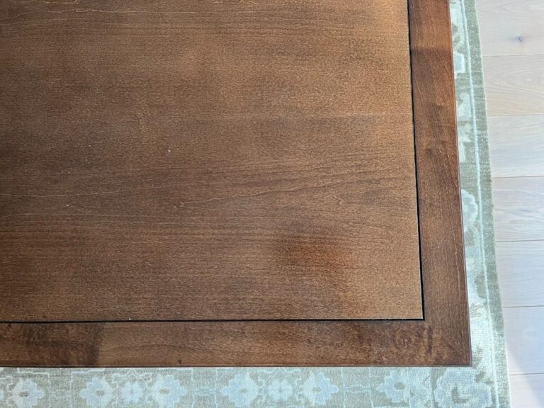 Wooden table with a square inset design, edged by a patterned green rug on a light wooden floor.