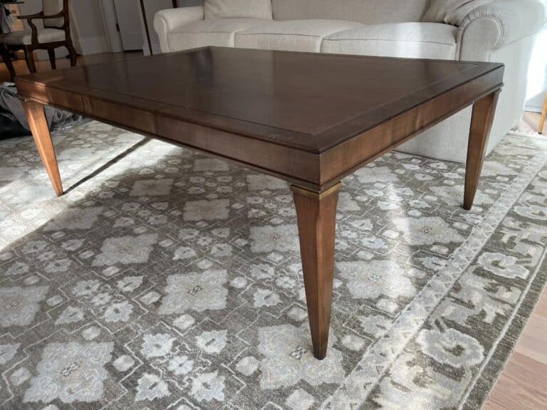 A wooden coffee table with tapered legs sits on a patterned rug in a living room with a beige sofa in the background.