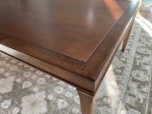 A wooden dining table with a dark finish is angled on a patterned rug, illuminated by sunlight.