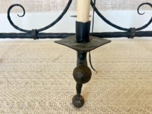 Close-up of a black wrought iron candle holder with a cream-colored candle on a textured surface. The design features ornamental curves and a round base.