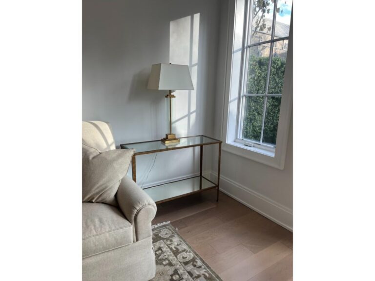 Room corner with a glass-top side table, brass lamp, beige sofa, large window, and patterned rug, illuminated by natural light.