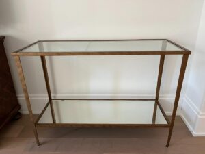 A gold-framed rectangular glass console table with two shelves, placed against a white wall on a wooden floor.