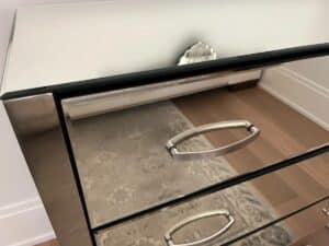 Close-up of a mirrored dresser with three silver handles, reflecting part of a rug and a decorative item near the back.