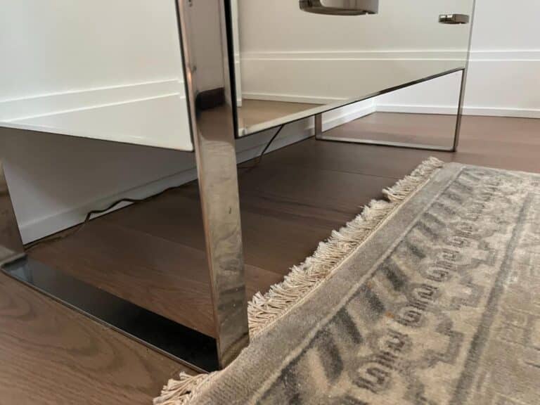 A reflective metal table leg stands on a wooden floor next to a patterned rug with fringe.