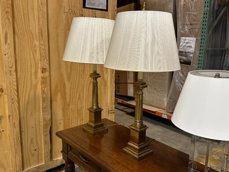 Two brass table lamps with white shades on a wooden table in a storage area with wooden panels and cardboard boxes in the background.