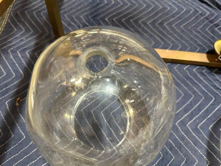 A clear glass bowl with a small circular opening rests on a blue zigzag-patterned fabric. Nearby, part of a metal frame is visible.