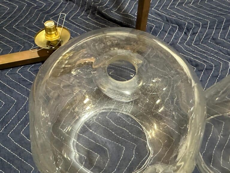 Close-up of a large, clear glass lampshade with a central opening, resting on a textured blue fabric. A lamp base with a bulb socket is partially visible in the background.