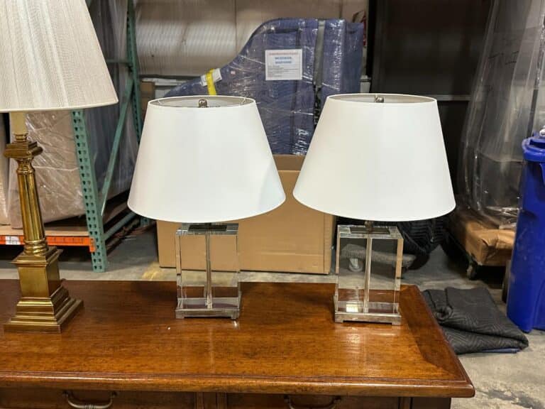 Two white lamps with square bases on a wooden table in a storage area, surrounded by boxes and wrapped furniture.