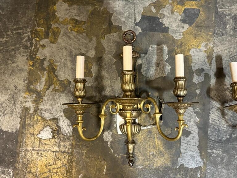 A brass three-candle wall sconce is mounted on a textured, decorative wall with gold and gray patterns.