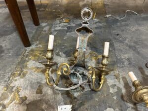 Antique brass chandelier with five candle-style holders and visible wear, lying on a textured floor beside a wooden table leg.