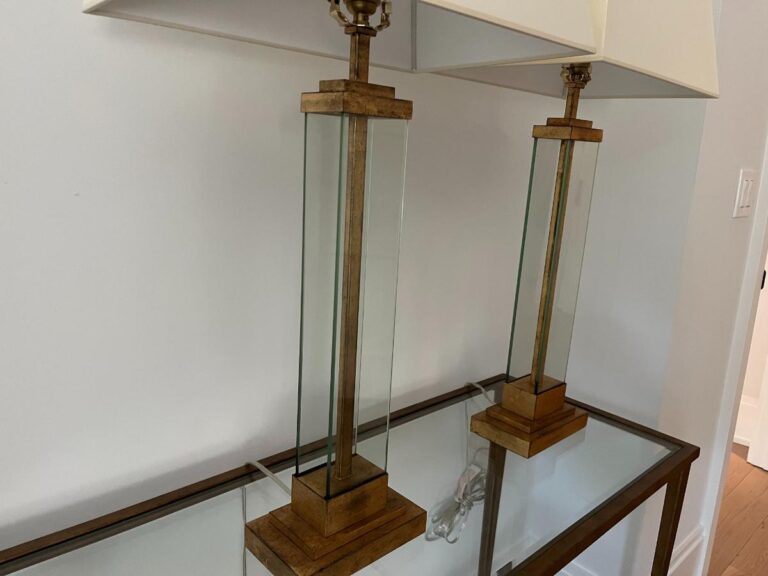 Two glass and wood table lamps with rectangular shades on a glass-top console table against a light-colored wall.