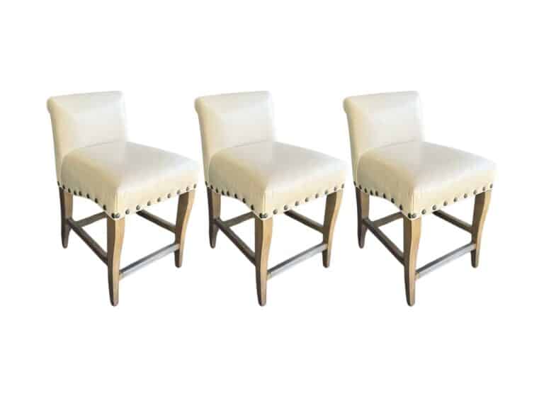 A set of three white leather counter stools with wooden legs and silver nailhead trims arranged in a row.